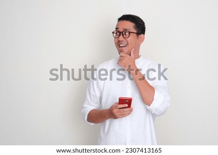 Adult Asian man holding phone and looking to the right side with amazed expression