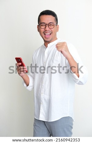Adult Asian man looking camera while holding mobile phone showing enthusiastic expression