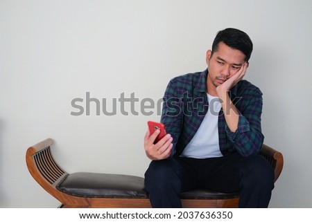 Adult Asian man sitting in a chair and showing boring gesture while looking to his mobile phone