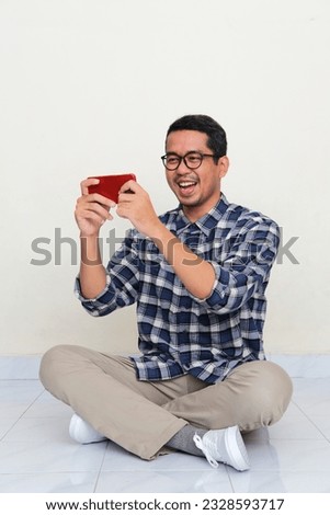 Adult Asian man sitting cross legged on the floor showing happiness when playing mobile games