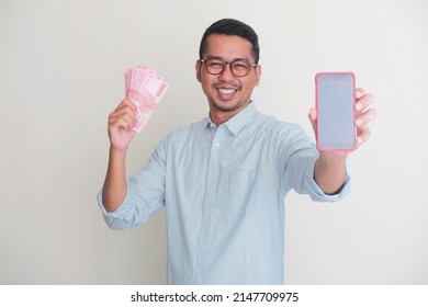 Adult Asian Man Smiling Happy While Showing Blank Mobile Phone Screen And Holding Paper Money