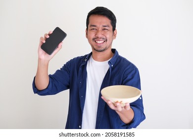 Adult Asian Man Smiling While Holding Empty Eating Bowl And Mobile Phone