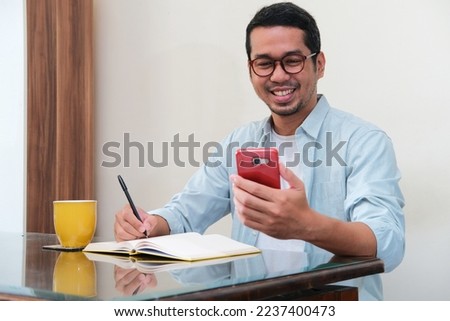 Adult Asian man smiling while looking to his handphone and write in a book