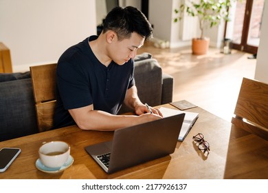 Adult Asian Man Taking Notes While Working  Studying In Cozy Livingroom At Home