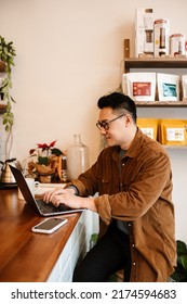 Adult asian man wearing eyeglasses smiling and using laptop in cafe indoors