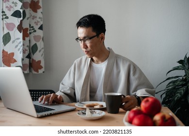 Adult Asian Man Wearing Glasses And Robe Sitting In Kitchen Using Laptop During Breakfast. Leisure Morning Time.