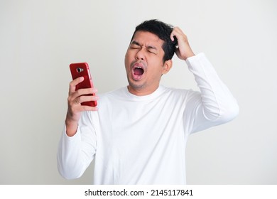 Adult Asian Man Yawning And Scratching His Head While Holding His Mobile Phone