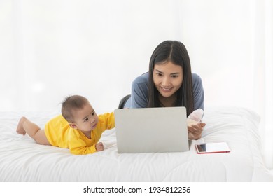 Adult Asian Woman Mom Smiling Do Feel Happy And Do Speaking On Smartphone And Look At Laptop With Baby Child On Bed In The Morning.