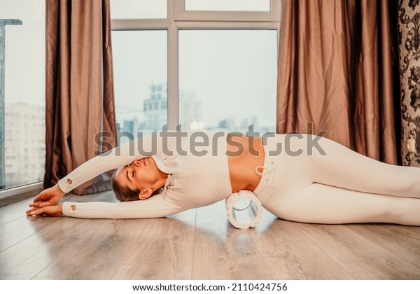 Adult athletic woman, in white bodysuit, performing\
fascia exercises on the floor - caucasian woman using a massage\
foam roller - a tool to relieve tension in the back and relieve\
muscle pain - the