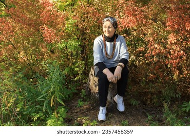 Adult attractive pensioner woman on a walk in the autumn park, stylish casual clothes,lifestyle, beautiful autumn background, yellow and red foliage,admiring nature,space for text,headdress, good mood - Powered by Shutterstock