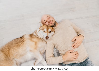 Adult Bald Man With Husky Puppy Sleeping On Floor. Owner With Pet Together At Home. Kind And Soulful Emotions. Lovely Dog Resting With Young Male. Guy With Beloved Domestic Animal Hugging Each Other.