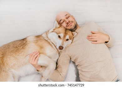 Adult Bald Man With Husky Puppy Sleeping On Floor. Owner With Pet Together At Home. Kind And Soulful Emotions. Lovely Dog Resting With Young Male. Guy With Beloved Domestic Animal Hugging Each Other.