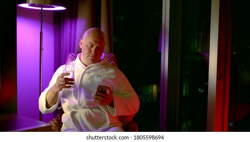 Adult Bald Man, Resting In A Chair After A Shower In A Bathrobe And Writing A Message On A Mobile Phone, Drinking Alcohol In The Evening.