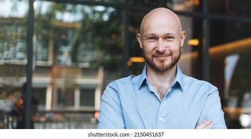 Adult bald smiling attractive man forty years with beard in blue shirt businessman against glass wall of street cafe, banner - Shutterstock ID 2150451637