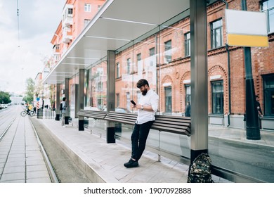 Adult bearded man with backpack standing on tram station and browsing smartphone on empty city street against brick buildings on cloudy day - Powered by Shutterstock