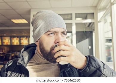 Adult bearded man in a beanie and a dark jacket drinks hot coffee in a cafe, melancholy - Powered by Shutterstock