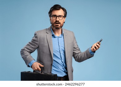 Adult bearded man in jacket and in glasses using smartphone and briefcase, posing in studio, isolated on blue. Business, technology, social media concept - Powered by Shutterstock