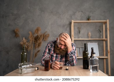 Adult Bearded Man Sitting At The Table Drinking A Bottle Of Alcohol At Home Sad Alone, Alcoholism, Holding His Head With His Hand. Signs And Symptoms Of Rehabilitation Abuse And Recovery Problems. 