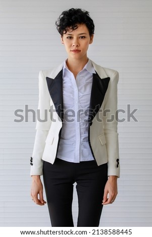 Adult beautiful fashionable Caucasian businesswoman with curly hairstyle wearing formal clothes with trendy suit, smiling with confidence and success, standing in indoor office and white background