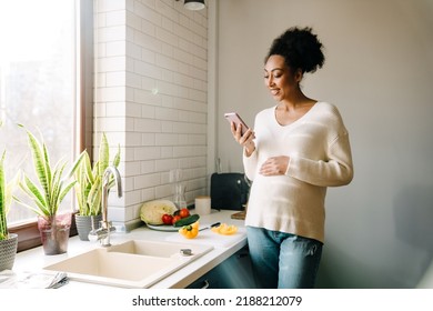 Adult beautiful smiling african pregnant woman looking on her phone and holding her belly while cooking salad in cozy kitchen at home - Powered by Shutterstock