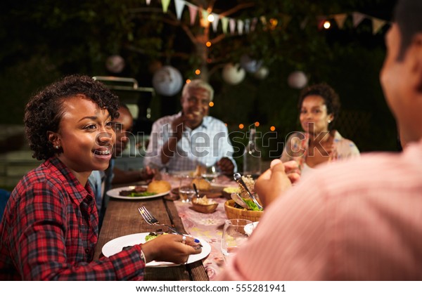 Adult black family eat dinner in garden, over\
shoulder view