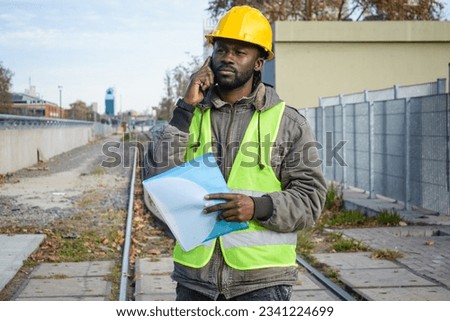 adult black man of african ethnicity maintenance builder engineer with safety helmet and reflective vest is talking on the phone standing on the railway tracks.