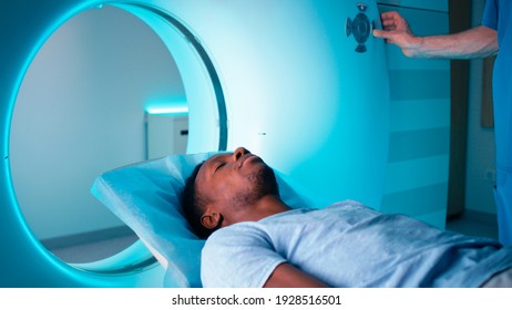 Adult Black Man Lying Down On Table Then Crop Doctor Pushing Button On CT Machine And Starting Scanning Procedure In Laboratory Of Clinic