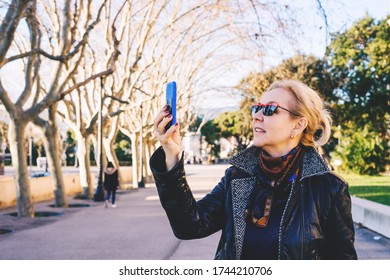 An Adult Blonde Lady Use Facetime To Talking On The Phone In A Dark Coat And Glasses Looks Up In Spring Park And Smiles. Side View.