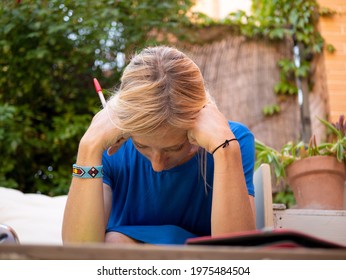 An adult blonde woman in a blue dress depressed hunched over her hands sitting in the garden with a wooden armchair and white cushions, with a wooden table that has a tablet, a mobile phone, sunglasse