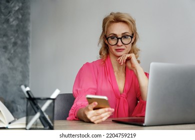Adult blonde woman in the office distracted from work by checking her social media accounts on the phone. Female sitting at her workspace with laptop and smartphone. Close up, copy space, background.