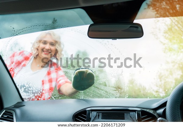 An adult blonde woman\
washes the windshield of a car with a washcloth, a view from inside\
the car interior