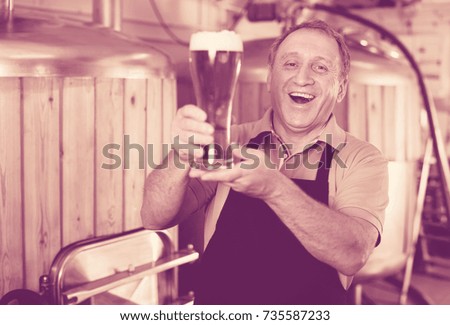 Adult brewer is standing with golden beer in glass in the fabric.