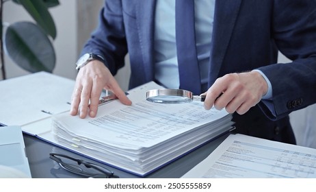 Adult businessman or auditor examining documents with magnifying glass. Professional in a suit scrutinizes financial reports, detail-focused work concept - Powered by Shutterstock