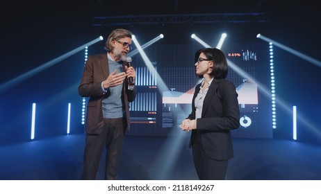 An Adult Businessman On The Stage In Casual Clothes Telling The Female Presenter And The Audience About The Graphs And Company Finance On The LED Screen In Studio, During A Business Training Lecture