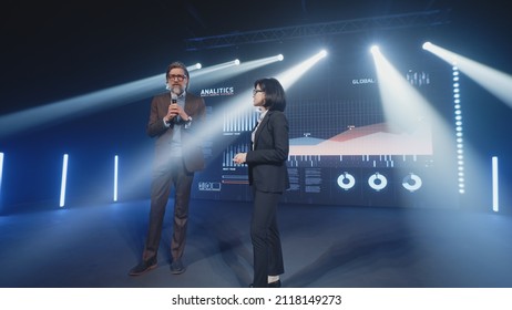 An Adult Businessman On The Stage In Casual Clothes Telling The Female Presenter And The Audience About The Graphs And Company Finance On The LED Screen In Studio, During A Business Training Lecture