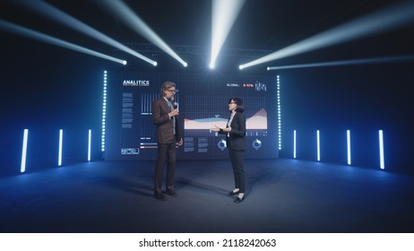 An Adult Businessman On The Stage In Casual Clothes Telling The Female Presenter And The Audience About The Graphs And Company Finance On The LED Screen In Studio, During A Business Training Lecture