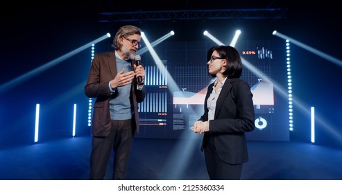 An Adult Businessman On The Stage In Casual Clothes Telling The Female Presenter And The Audience About The Graphs And Company Finance On The LED Screen In Studio, During A Business Training Lecture