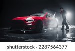 Adult Car Detailer in Uniform Washing a Red Sportscar with a High Pressure Cleaner. Cleaning Technician Working on a Stylish American Car in a Dark Room. Commercial Studio Photo for Advertising