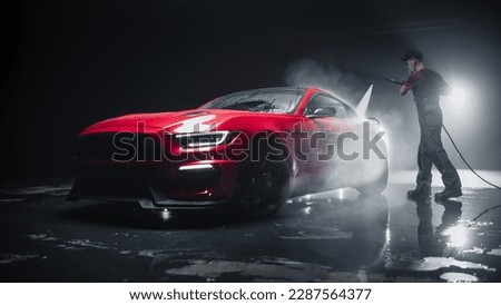 Adult Car Detailer in Uniform Washing a Red Sportscar with a High Pressure Cleaner. Cleaning Technician Working on a Stylish American Car in a Dark Room. Commercial Studio Photo for Advertising