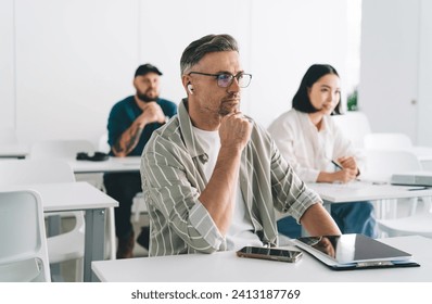 Adult Caucasian male in bluetooth earbuds and optical spectacles for vision correction listening audio book during learning cooperation in university, businessman with colleagues on office interior - Powered by Shutterstock