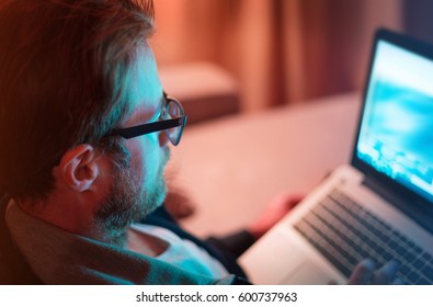 Adult caucasian man in glasses and casual clothes working by night on laptop computer - close up. Home, living room interior as background. Freelance worker or leisure (after hours) concept.