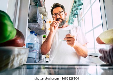 Adult Caucasian Man Taking Note List Of Food Looking Inside The Open Fridge At Home - Kitchen Activity And Alternative Point Of View - Market Shopping And Quarantine Lockdown Stayhome Coronavirus Time