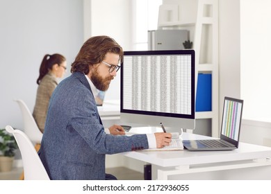Adult Caucasian man works as accountant or auditor sits at table with monitor and laptop, fills out tables and makes entries in workbook calculating profits and taxes located in open space office - Shutterstock ID 2172936055