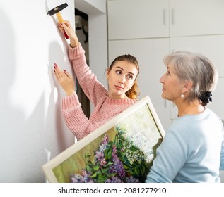 Adult caucasian woman helping her senior mother to put painting on wall. Daughter hammering nail, her mother standing beside with picture in hands.