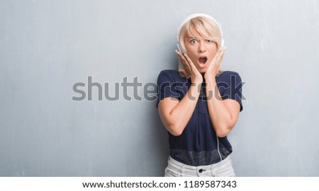 Adult caucasian woman over grunge grey wall wearing headphones scared in shock with a surprise face, afraid and excited with fear expression