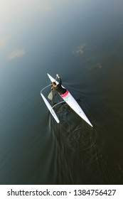 Adult Caucasian woman viewed from above and behind paddling in white outrigger canoe with float on dark water background with copy space