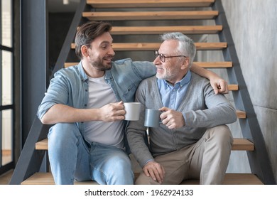 Adult Caucasian Young Son Supporting And Hugging Embracing His Old Senior Elderly Father Drinking Tea Together Sitting On The Stairs At Family Home. Happy Father`s Day!