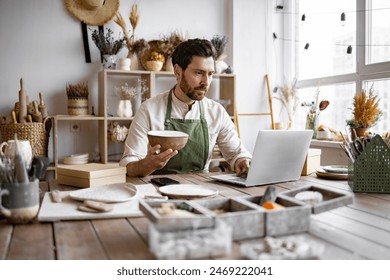 Adult ceramic shop owner using laptop while working in store. Male entrepreneur managing online order of mug on website. Confident young businessman running creative small business. - Powered by Shutterstock