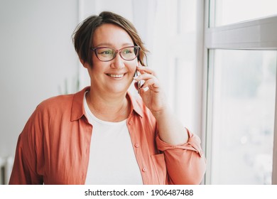Adult charming brunette woman businesswoman in the glasses plus size body positive using mobile near window