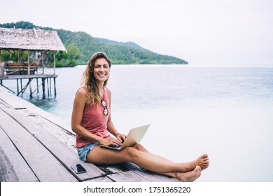 Adult cheerful pretty female in light summer clothes smiling brightly and looking at camera while sitting and using digital device on wooden pier near smartphone on nature